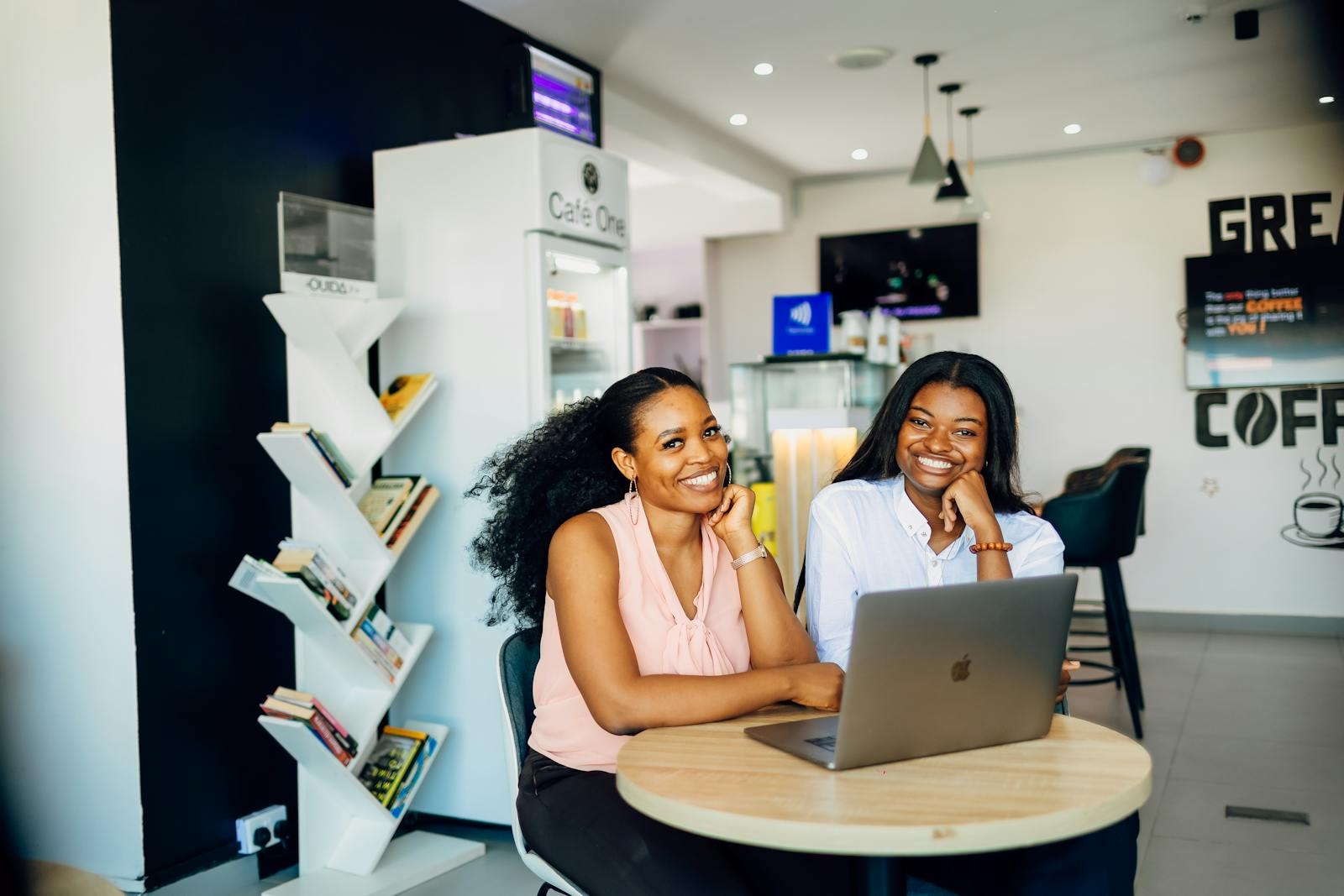 Two smiling businesswomen collaborate over a laptop in a vibrant Lagos café.