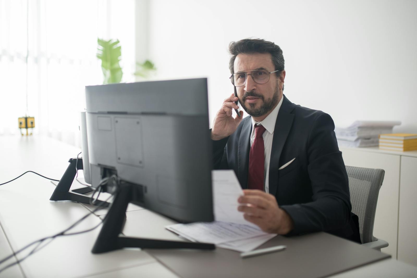 Confident businessman in a black suit making a phone call while reviewing documents in a modern office setting.