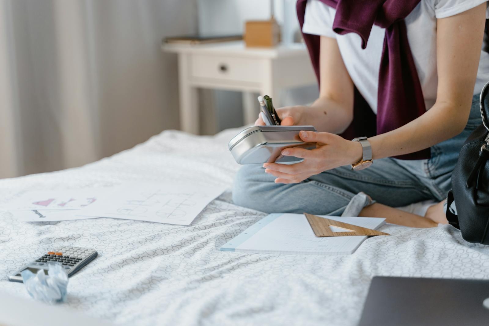 A student sitting on a bed while organizing study materials in a cozy bedroom environment.