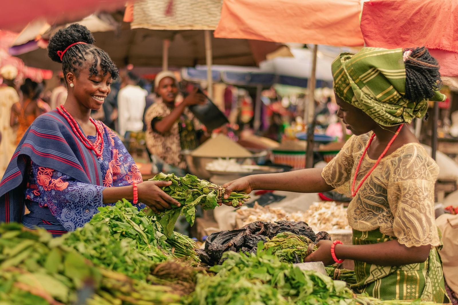 Two women in traditional attire exchange greens at a lively outdoor market. Vibrant and cultural.