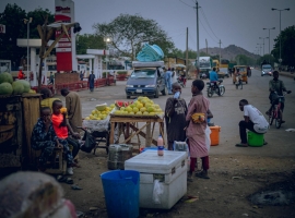 Vibrant street market in the evening with local vendors selling fruits and commuters passing by.