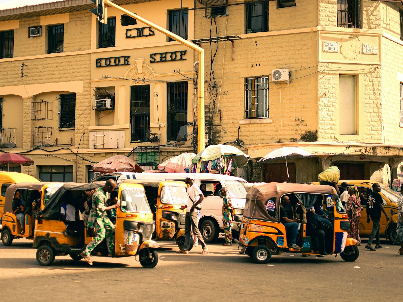 Vibrant city intersection with colorful auto rickshaws and pedestrians in front of a bookshop.