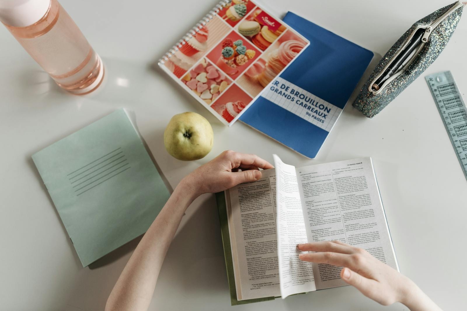 Overhead view of hands turning book pages at a study desk with notebooks, apple, and water bottle.